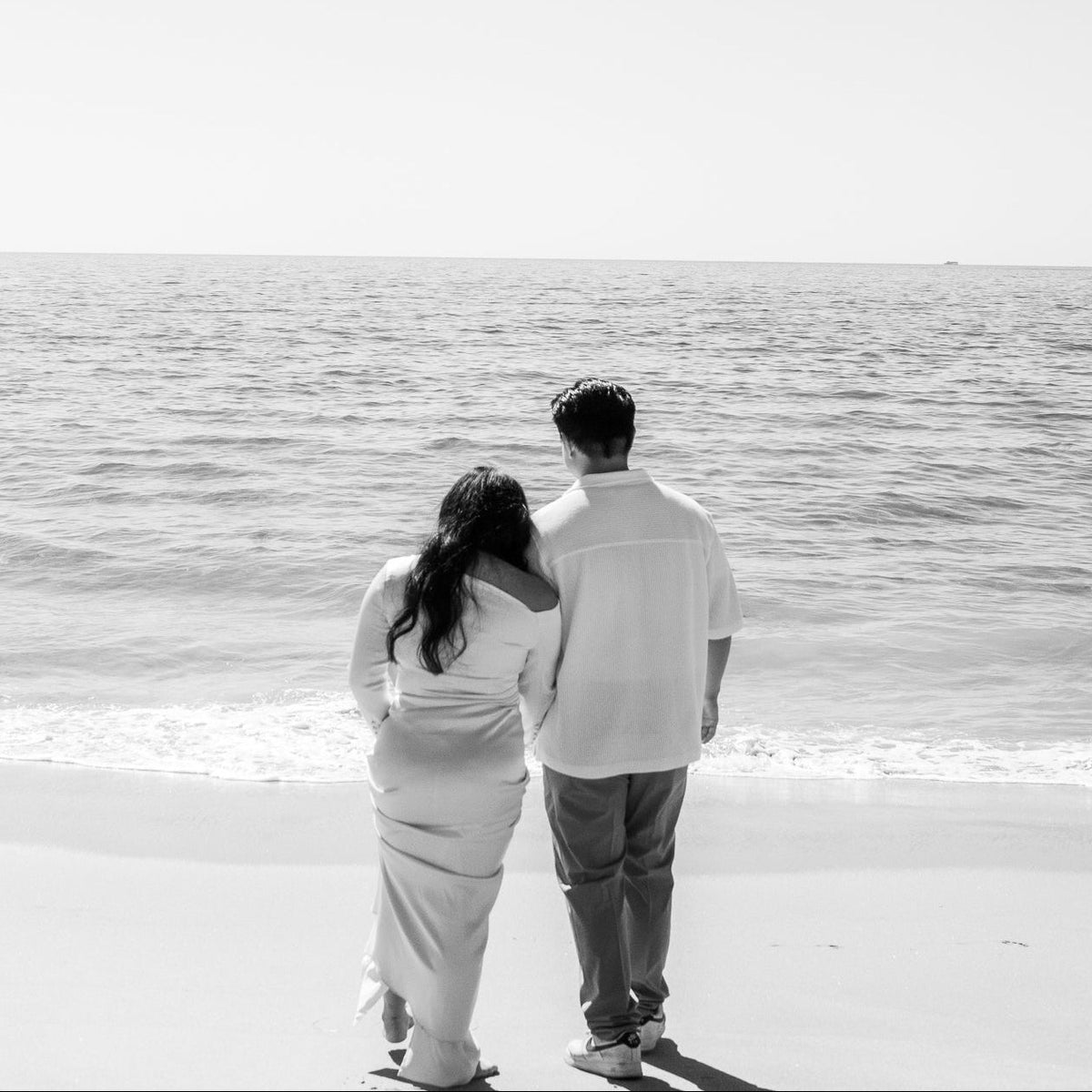 A black and white photograph of a couple from behind, standing on a sandy beach and looking out toward the ocean under a clear sky.