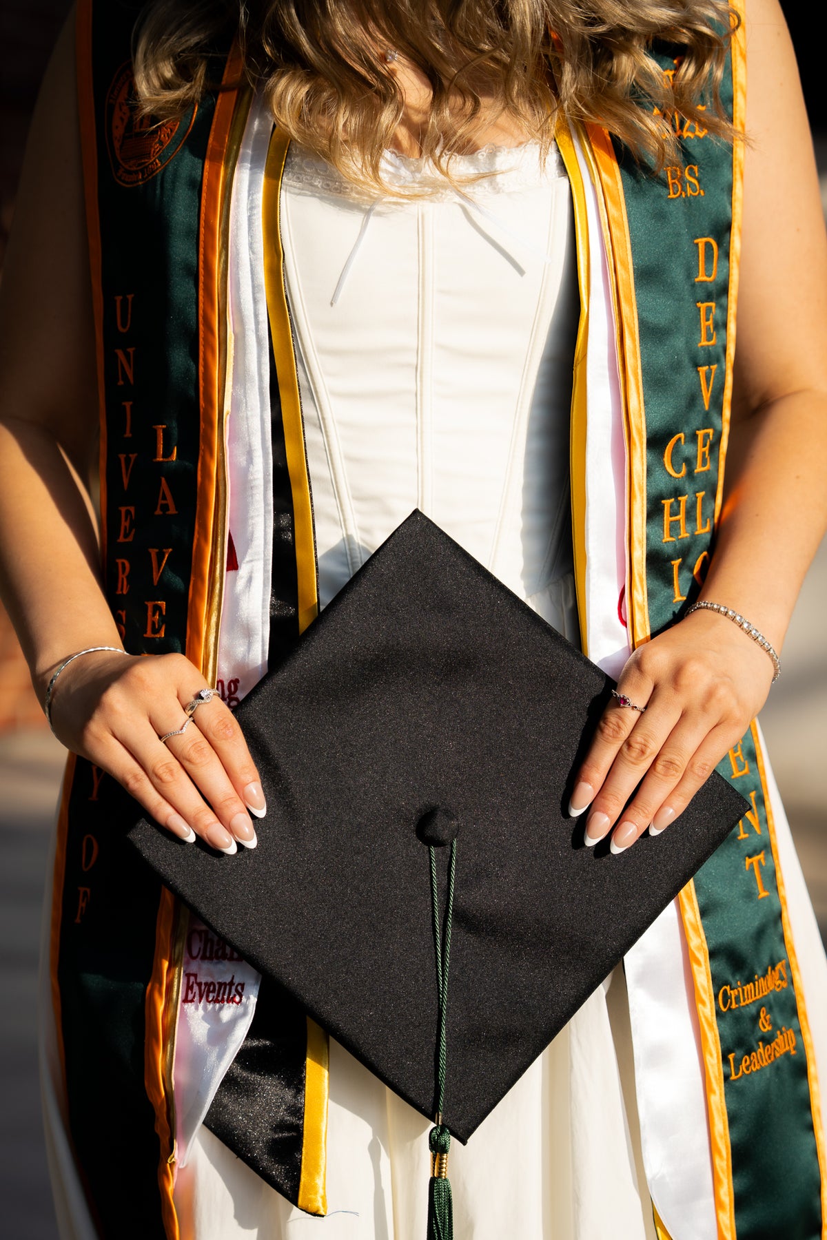 A close-up of a person holding a black graduation cap with a tassel, wearing a white gown and a green academic cap gown with gold and white stripes.